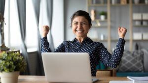 Eufórico joven estudiante india ganadora celebrar victoria triunfo sentarse en casa escritorio con computadora portátil ganar fortuna en línea sentirse emocionado obtener nueva oportunidad de trabajo buen examen resultado gran concepto de noticias.