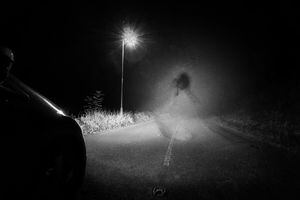 A spooky ghost of a woman in the road below a single street light in the countryside. Lit up by car headlights at night. With a grunge, vintage, old edit