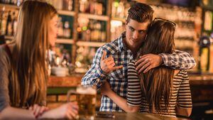 Joven infiel en un café. Está abrazando a su novia y haciendo un gesto de llamada a otra mujer a sus espaldas.