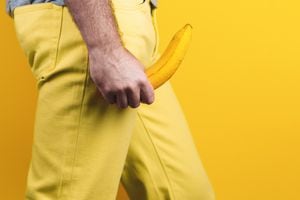 Potency and men's health. A man in yellow jeans holds a banana in his hands, at the level of the genitals, personifying an erection. Yellow background. Close up. Copy space. Side view.