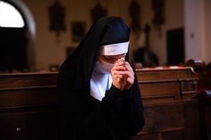 Nun holding hands together, praying to God, unrecognizable face. She is wearing a white surgical face mask.