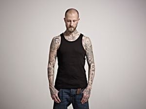 Portrait of skinny bearded male with heavily tattooed arms wearing a black vest top and jeans, looking to floor with his thumbs in his pockets.