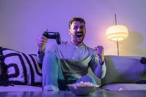 Young handsome man holding PlayStation joystick and cheerfully shouting . He sits on sofa in the living room and having great time with friends playing games online