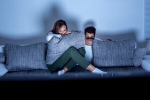 Young smiling couple watching a scary movie at home. Both Caucasian, casual, about 20 years old.