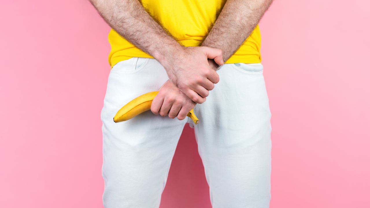 Potency and men's health. A man in white jeans, legs apart, holds a banana near the genitals. Pink background. Close up of hands.