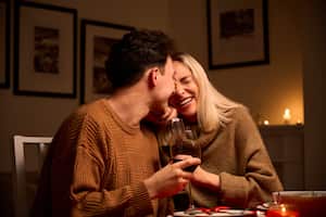 Happy young couple in love hugging, laughing, drinking wine, enjoying talking, having fun together celebrating Valentines day dining at home, having romantic dinner date with candles sitting at table.