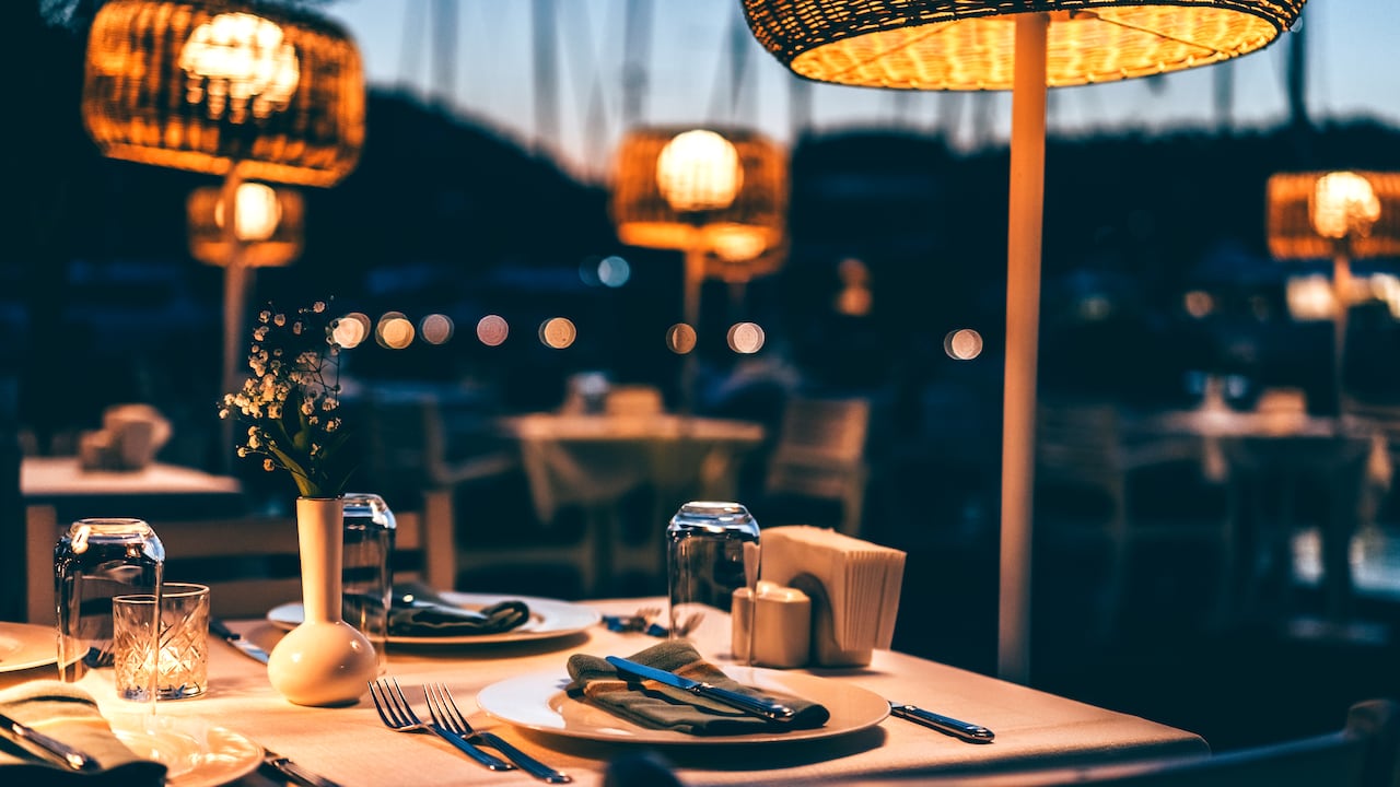 An elegant and romantic dinner setup for a night out, with no people in the background, the abstract design setup at twilight. The tables, elegantly laid out, and woven basket pendant lights provide a cozy and inviting atmosphere. A serene and peaceful environment, perfect for a chic restaurant experience by the waterfront, with the gentle breeze and distant chatter filling the air.