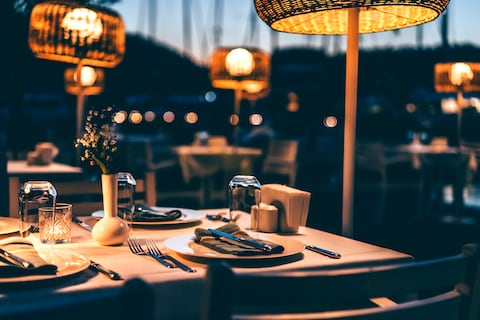 An elegant and romantic dinner setup for a night out, with no people in the background, the abstract design setup at twilight. The tables, elegantly laid out, and woven basket pendant lights provide a cozy and inviting atmosphere. A serene and peaceful environment, perfect for a chic restaurant experience by the waterfront, with the gentle breeze and distant chatter filling the air.