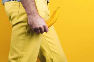 Potency and men's health. A man in yellow jeans holds a banana in his hands, at the level of the genitals, personifying an erection. Yellow background. Close up. Copy space. Side view.