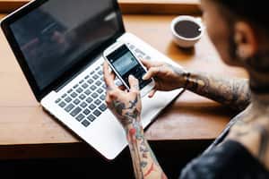 A close up shot of a young tattooed woman's hands  checking her smart phone in a café while working on her Laptop. The room is bright and there' a cup of coffee in front of her.