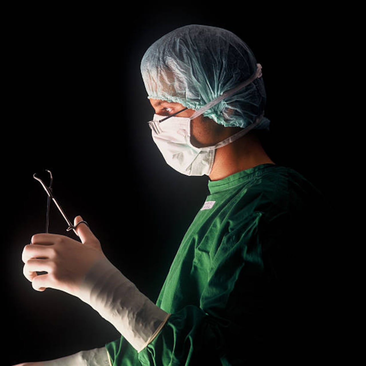 Surgeon or doctor, performing a surgical procedure or operation or surgery with a medical clamp, wearing a medical mask and special glasses, black background (Tuchklemme nach Backhaus)
