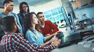 Primer plano de un grupo de desarrolladores de aplicaciones discutiendo sobre cierto código para una aplicación. Hay tres hombres y dos mujeres de pie y sentados frente a una computadora. Disparo inclinado.