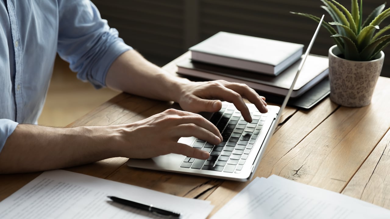 Hombre de negocios de cerca usando una computadora portátil, escribiendo en el teclado, sentado en un escritorio de madera con documentos, escribiendo un correo electrónico, contable escribiendo un informe financiero, estudiante ocupado estudiando en línea, buscando información