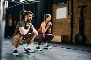 Young athlete using kettlebells while exercising squats during sports training in health club. Focus is on man.