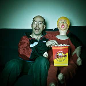 Nerdy looking guy and blow up doll watching TV