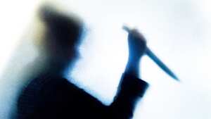 Backlit image of the silhouette of a person, probably a woman, wielding a sharp knife in an aggressive way. The silhouette is distorted, and the arms elongated, giving an alien-like quality. The image is sinister and foreboding, with an element of horror. The image conveys a domestic violence, knife crime theme. Horizontal image with copy space.