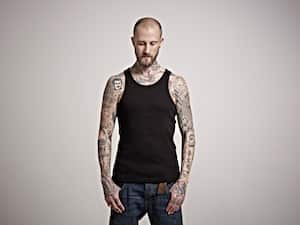 Portrait of skinny bearded male with heavily tattooed arms wearing a black vest top and jeans, looking to floor with his thumbs in his pockets.