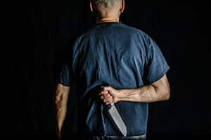 Man holding kitchen knife in his back, wearing black t-shirt, on black background