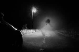 A spooky ghost of a woman in the road below a single street light in the countryside. Lit up by car headlights at night. With a grunge, vintage, old edit