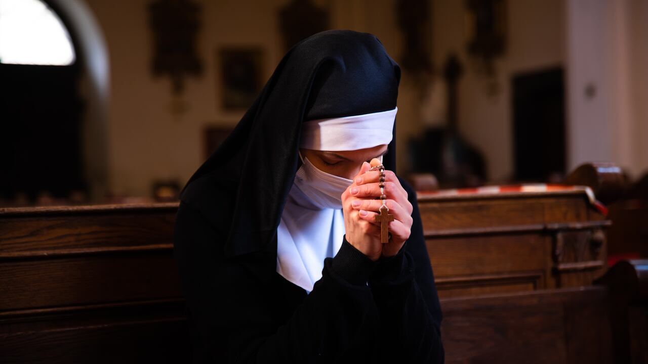 Nun holding hands together, praying to God, unrecognizable face. She is wearing a white surgical face mask.